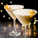 Two martini glasses filled with a frozen orange and coconut cocktail on a black background with twinkle lights
