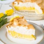 A lemon pie slice with a full pie in the background on a white table