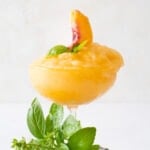Peach Bellini recipe in a wide mouth champagne glass decorated with peach slices and basil.