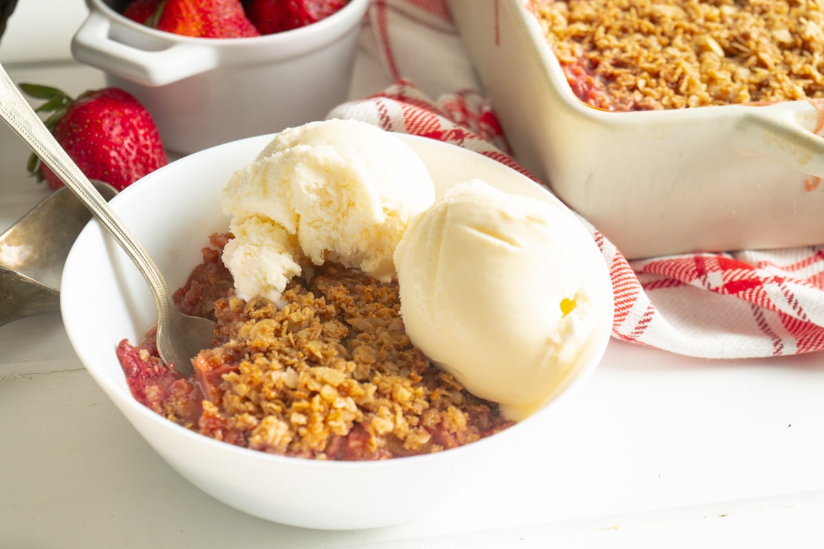 Strawberry rhubarb crisp in a white bowl with ice cream on a table with the strawberry rhubarb dessert dish nearby.