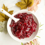 A small bowl of Orange Cranberry Sauce with a white spoon surrounded by fall leaves.