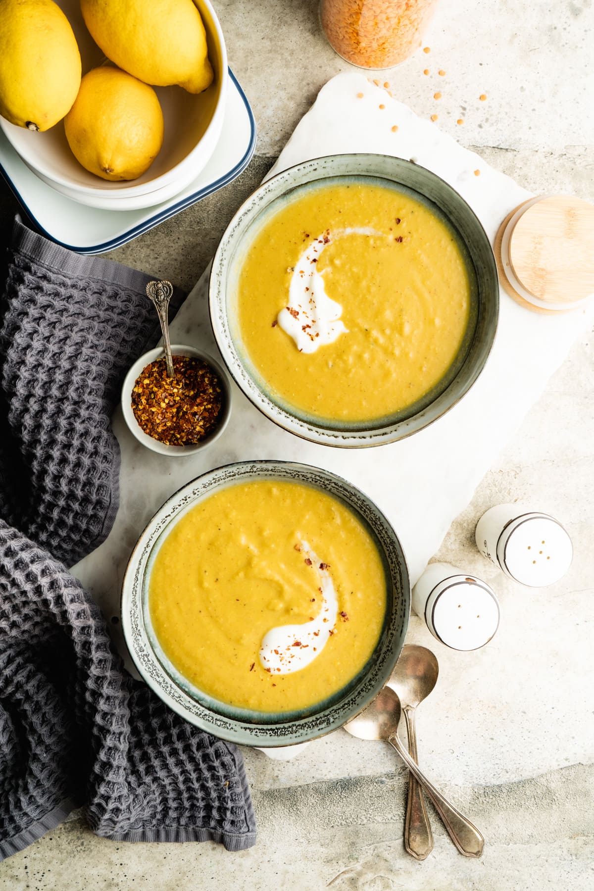 Two bowls of spicy red lentil soup on a table with chili flakes and lemons.