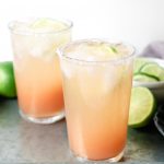 Two highball glasses rimmed with salt and filled with a paloma cocktail on a tray with limes