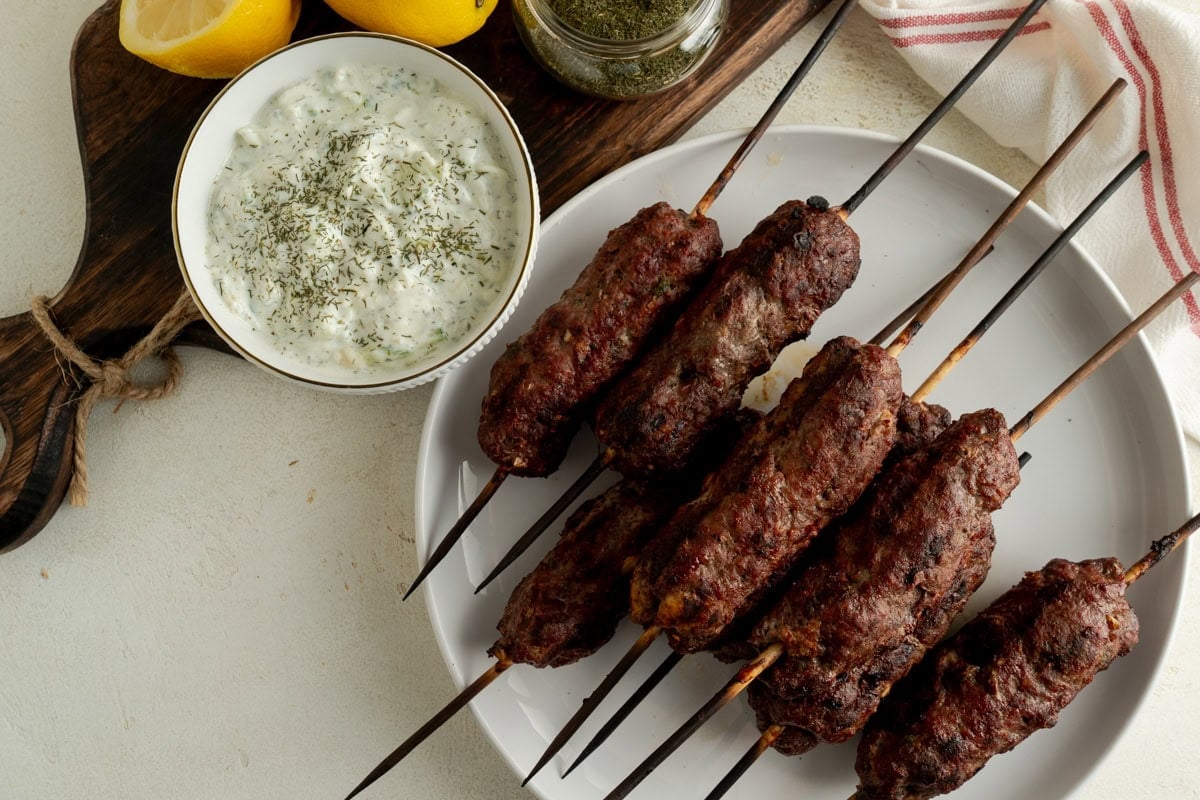A plate of kofta kebobs beside a bowl of tzatziki sauce.