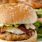 A close up of a chicken burger dressed with lettuce, bacon and mayo on a bun with more in the background