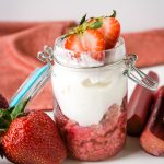 A jar of heatlhy strawberry overnight oatmeal with strawberries and rhubarb beside the jar on a white table with a red cloth napikin