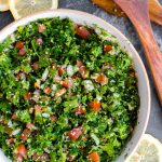 A large bowl of tabbouleh salad on a table with salad tongs and lemons