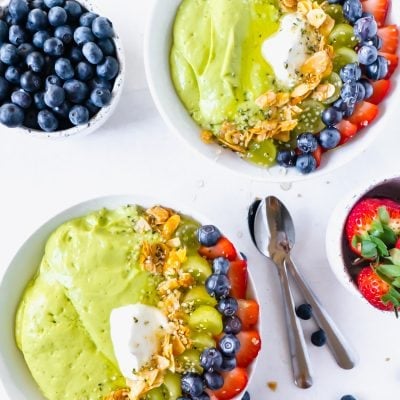 Two smoothie bowls on a white table surrounded by fruit