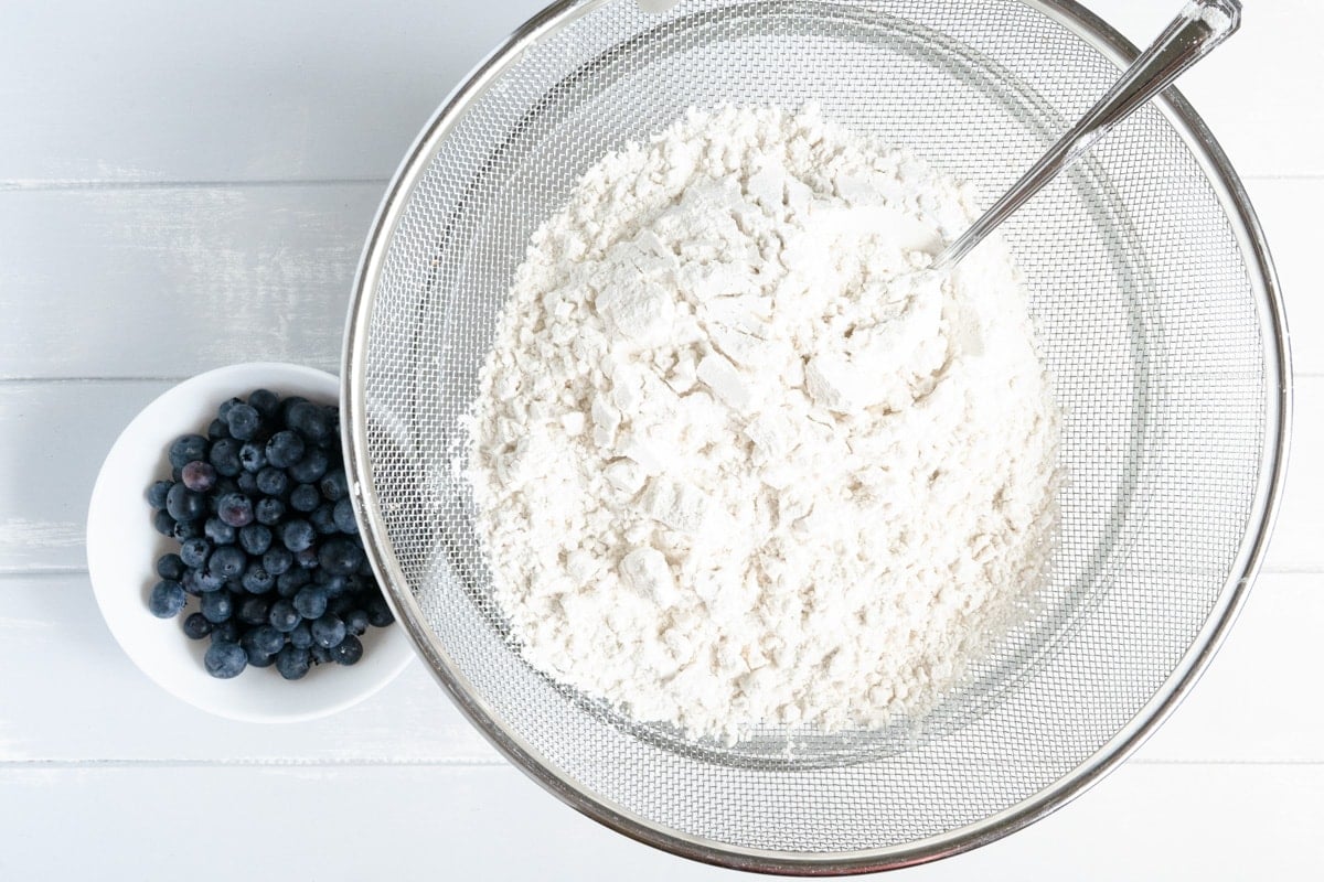 A bowl sieve filled with flour over a bowl with a smaller bowl of blueberries beside.