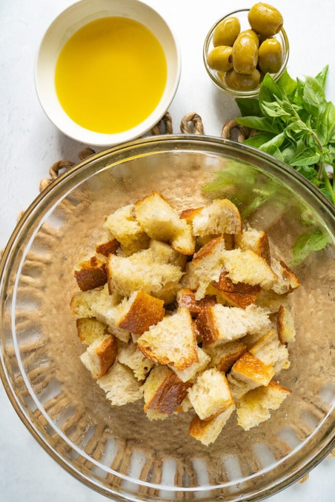 Cubed bread in bowl, next to olive oil, olives and basil. 