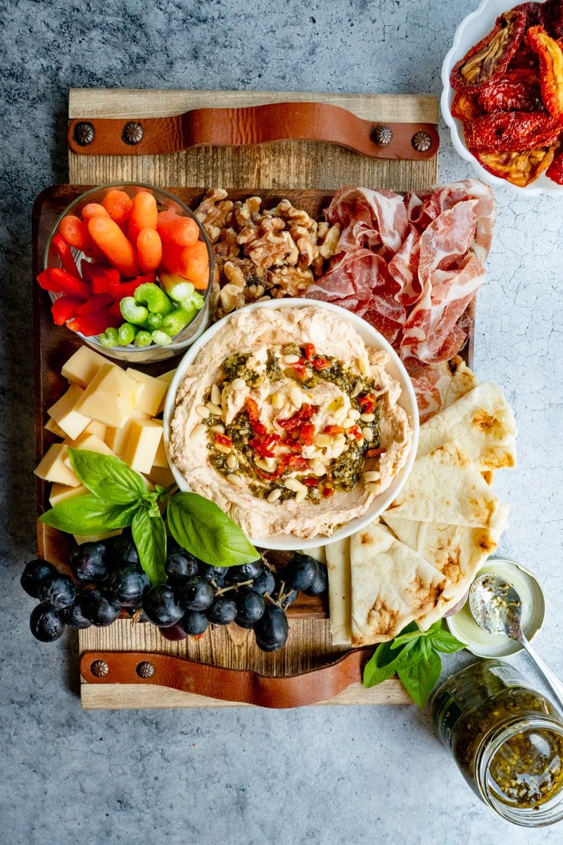 A charcuterie board with a pesto cream cheese dip recipe in the center.