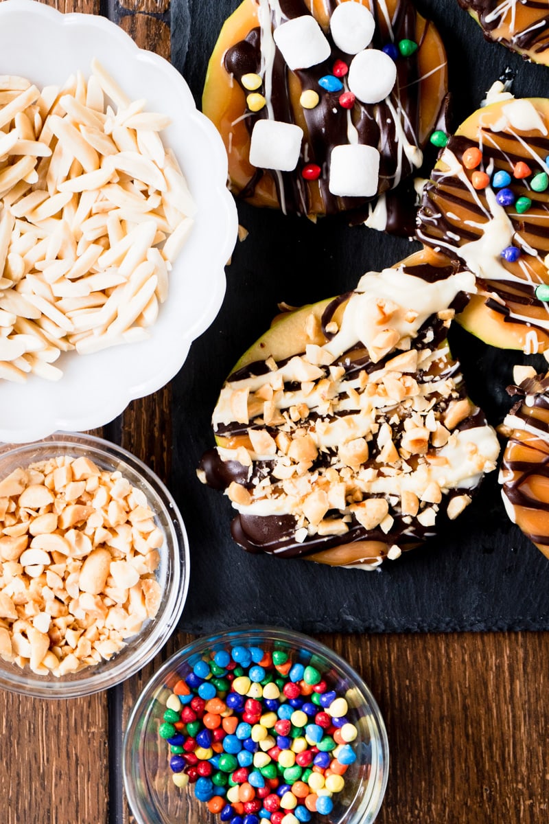 Chocolate covered apple slices being decorated with nuts and sprinkles.