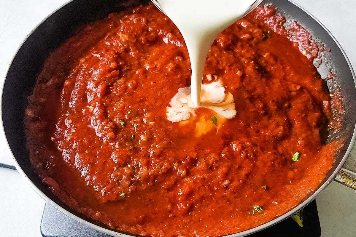 Heavy cream being poured into a pan of marinara sauce to make a vodka sauce.