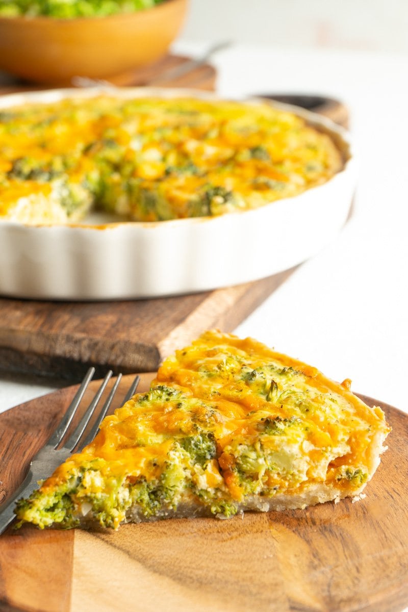 A slice of a breakfast quiche made with broccoli and cheddar on a wood plate with a fork.