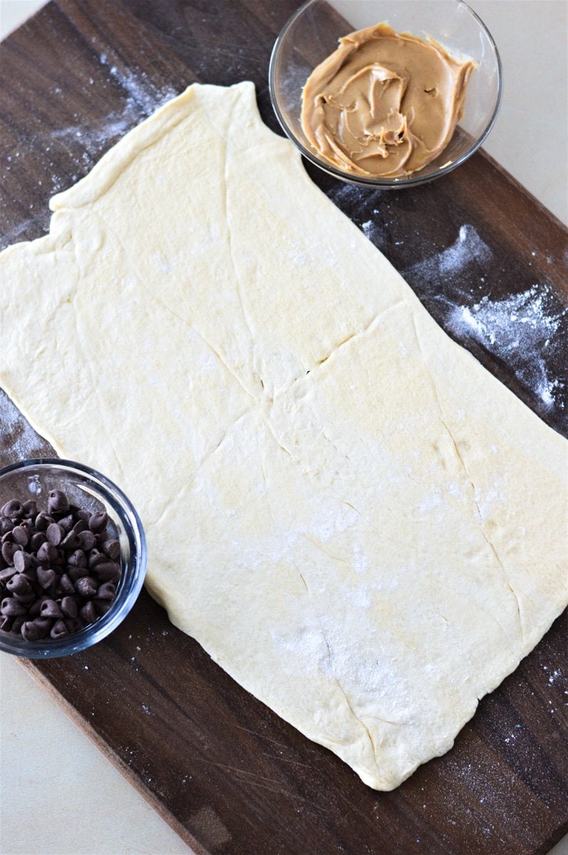 Crescent roll dough rolled out and sealed on a cutting board with a bowl of chocolate chips and peanut butter.