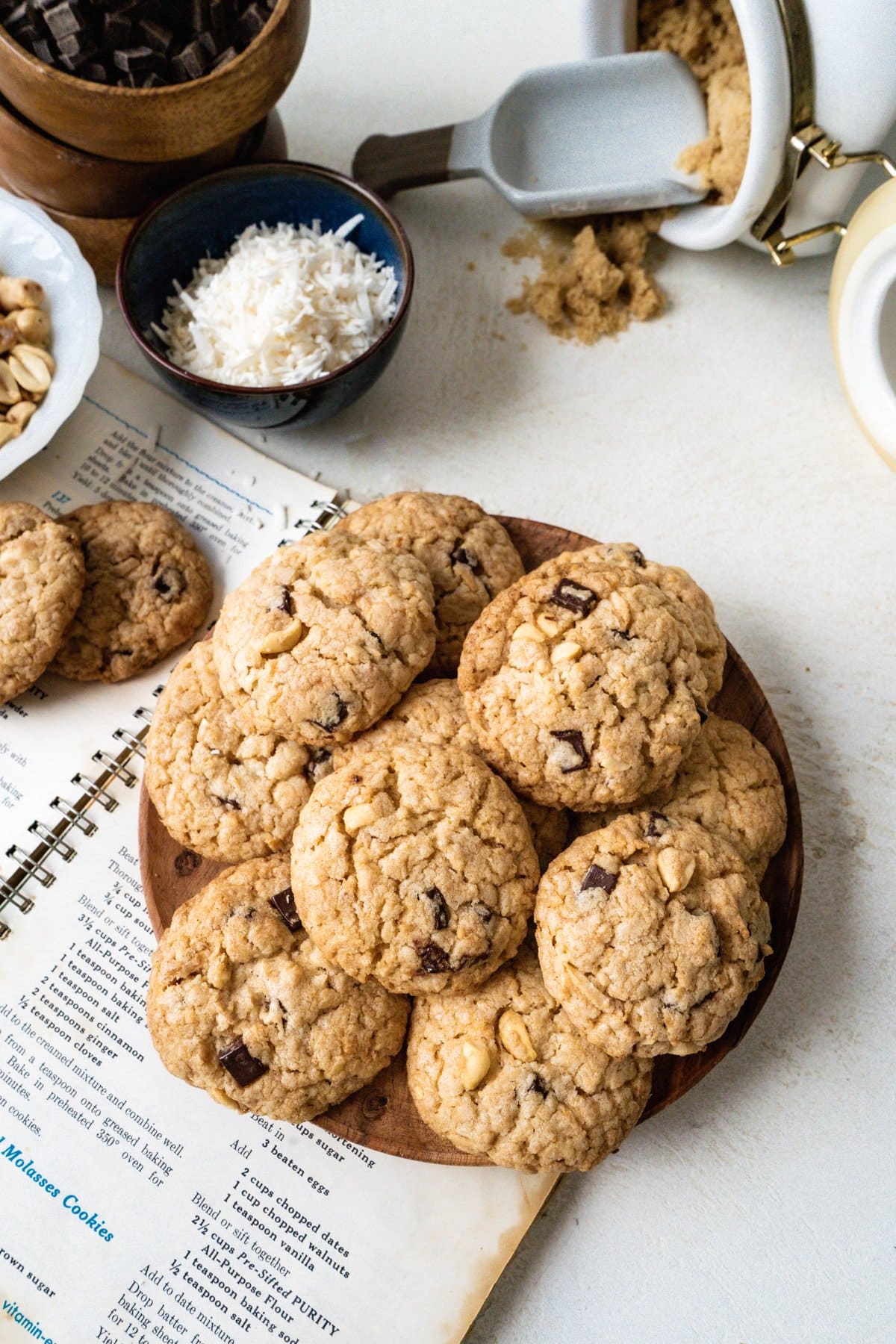 Chocolate oatmeal cookies made with coconut and salted peanuts on a wood plate on top a a table with baking supplies and a cookbook.