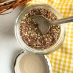 A small jar of a zaatar spice blend with a spoon on a yellow checkered napkin.