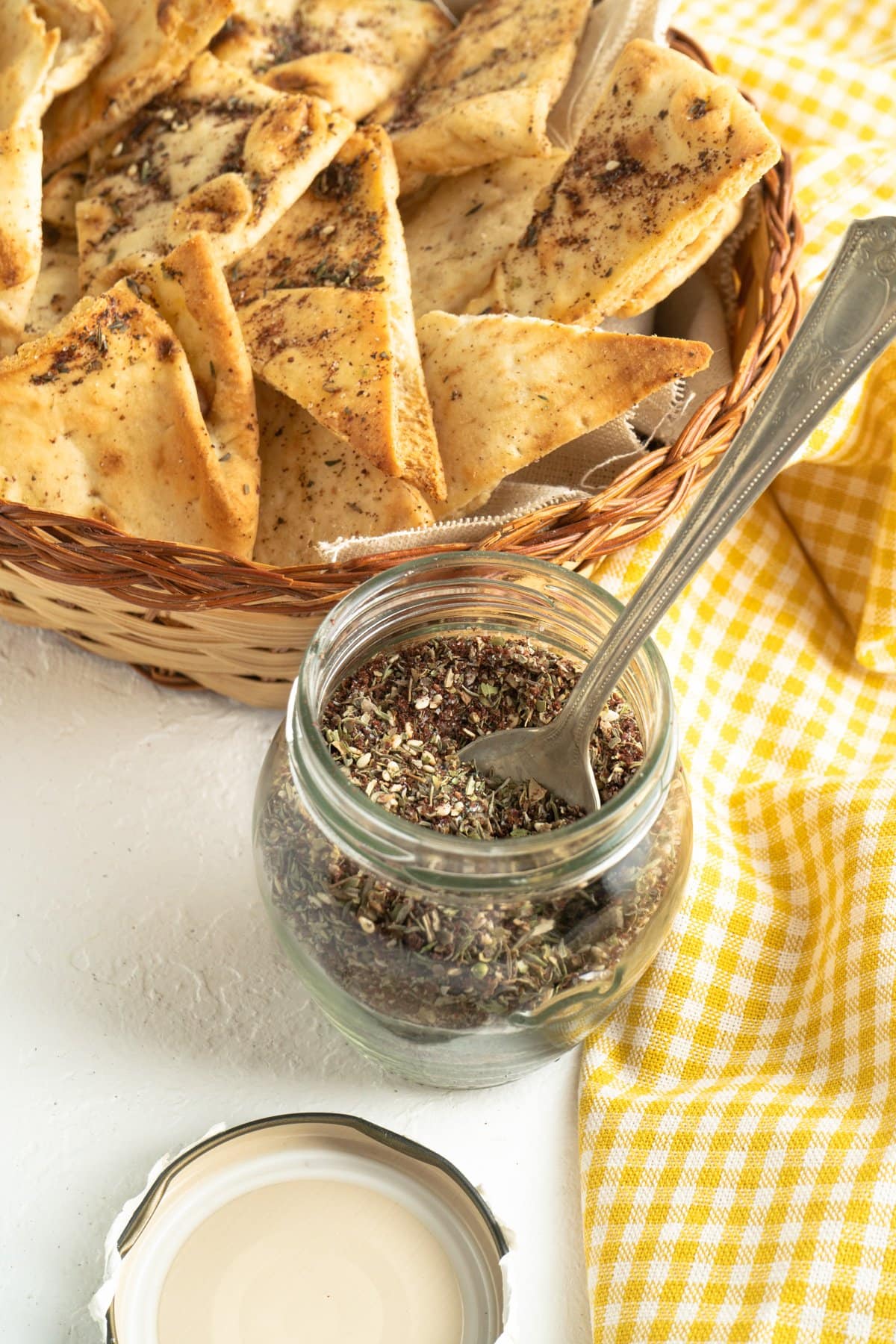 A jar of zaatar spice with a spoon near some pita chips.