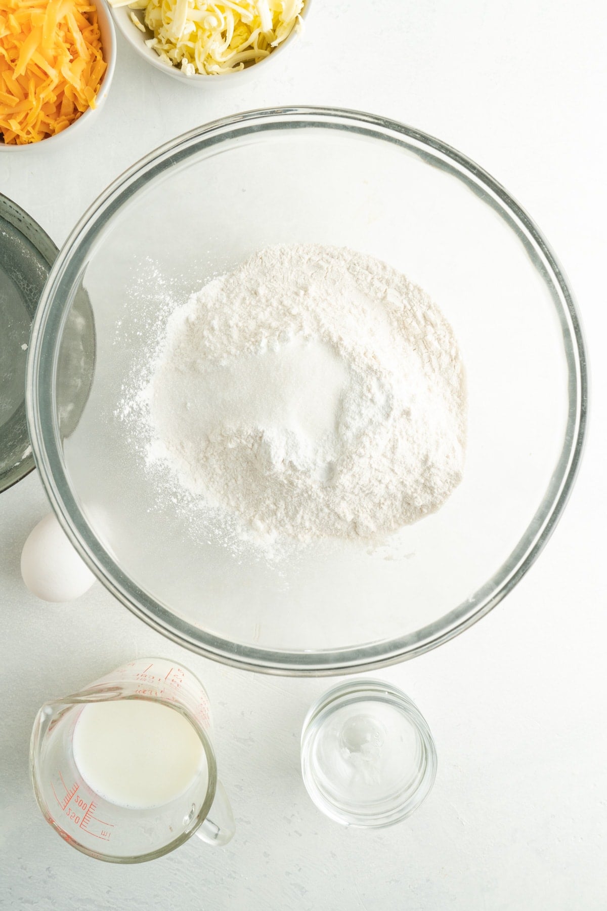 Glass bowl with flour in it and ingredients in measuring cups around it.