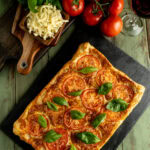 A tomato puff pastry tart recipe on a table with ingredients and a glass of red wine.