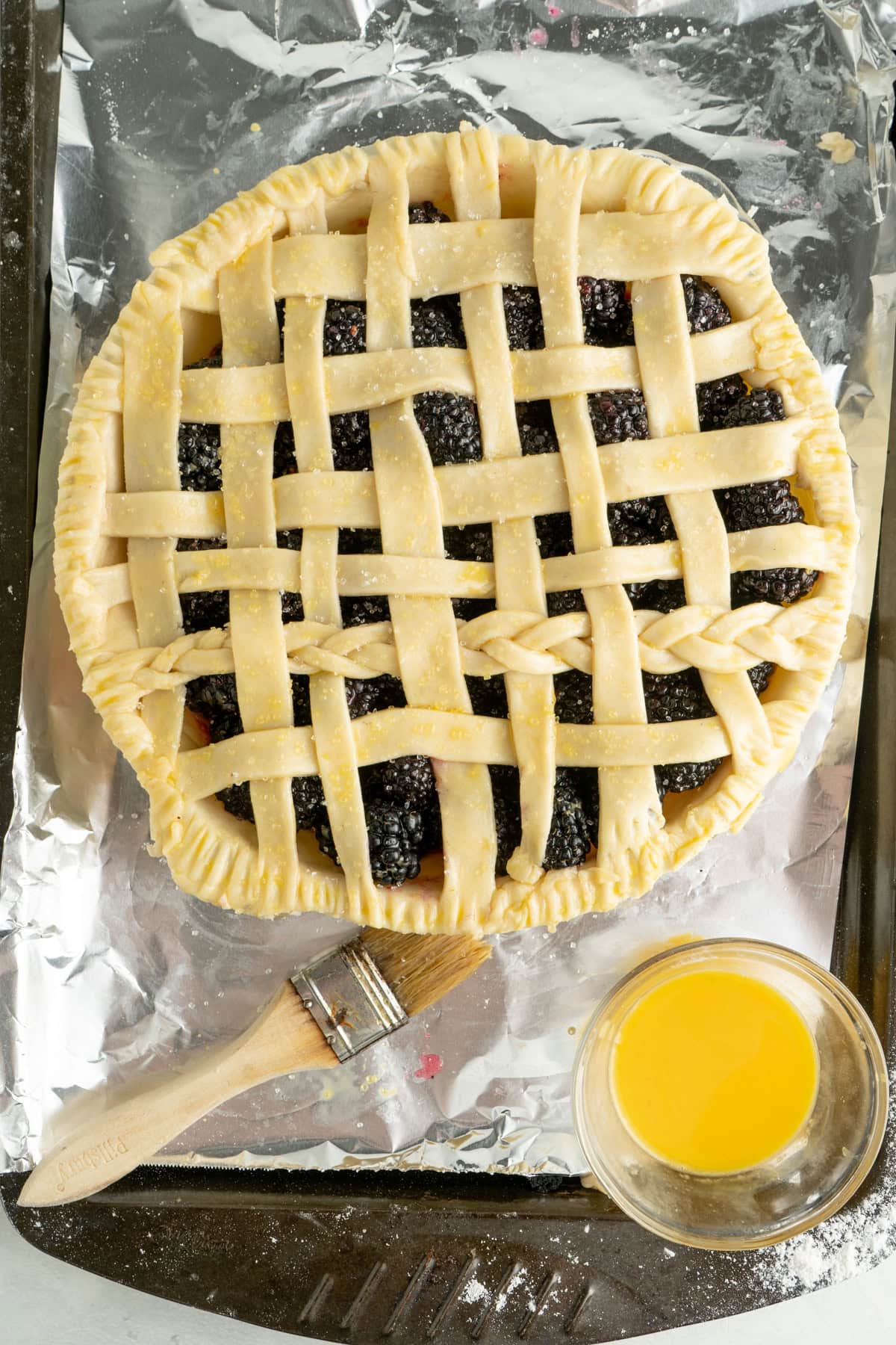 A blackberry pie recipe on a foiled baking sheet with a woven top being egg washed.