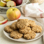 A plate of oatmeal applesauce cookies on a table with apples.