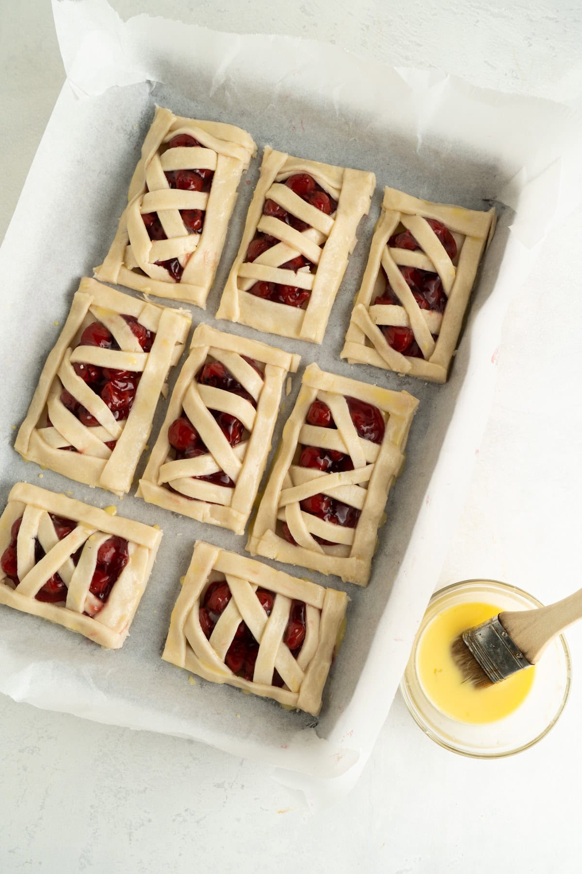 A cherry handpie recipe on a baking sheet being basted with an egg wash.