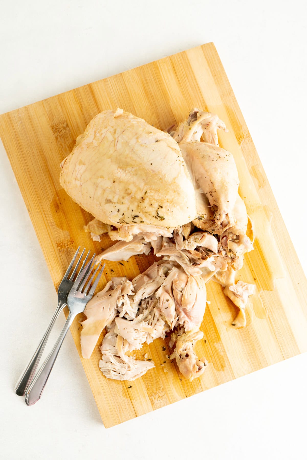 A whole boiled chicken on a cutting board being shredded with forks.
