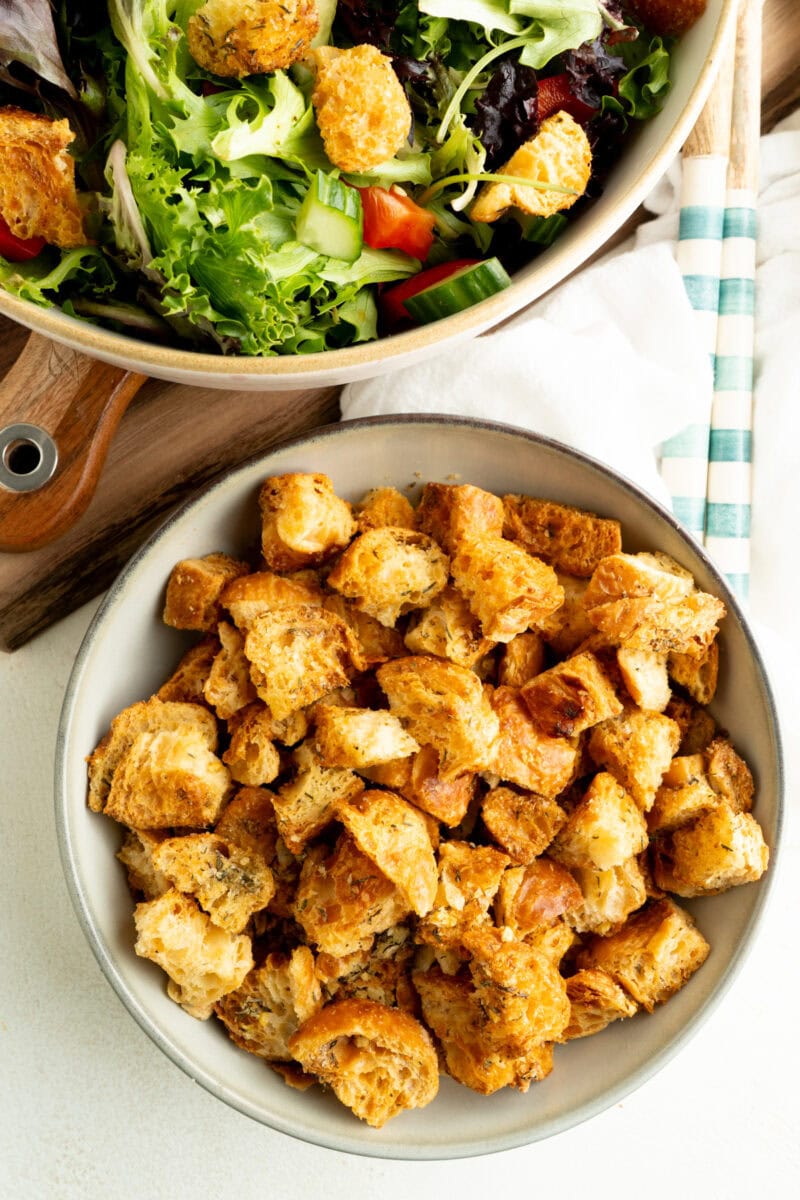 A bowl of croissant croutons with a salad nearby on a table.