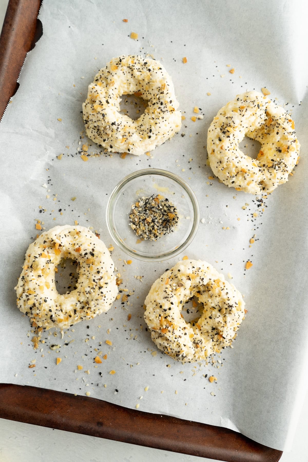 Unbaked bagels being sprinkled with everything bagel seasoning.
