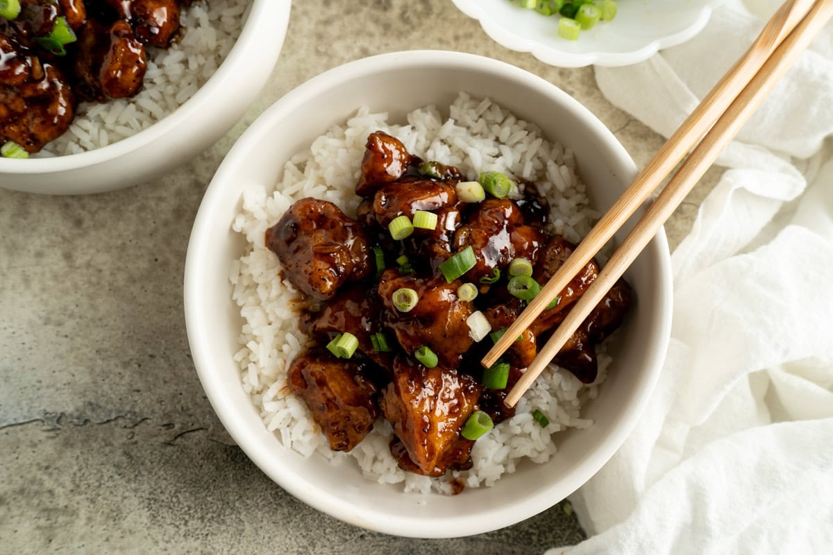 A bowl of honey pepper chicken over white rice with some green onions sprinkled on top.