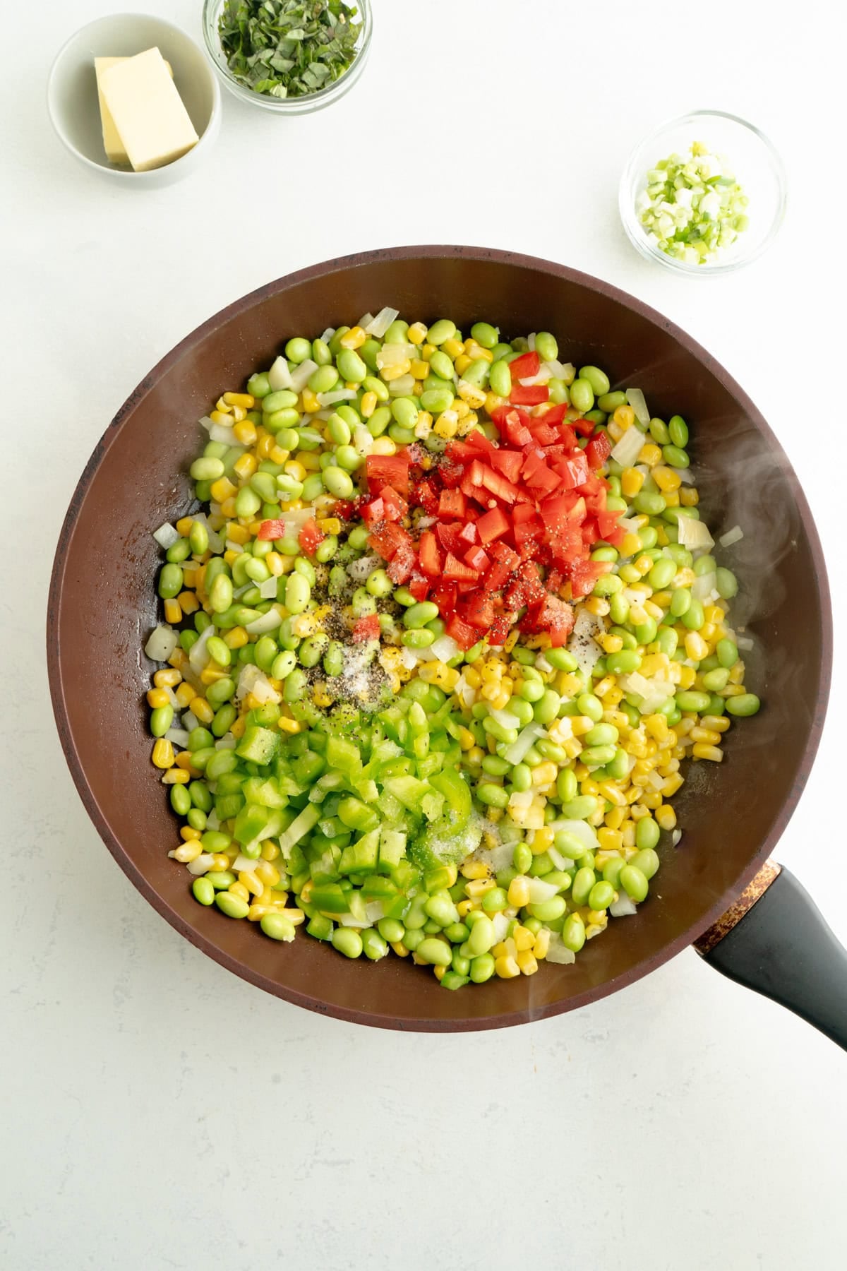 Red and green peppers being added to a pan full of corn and edamame.