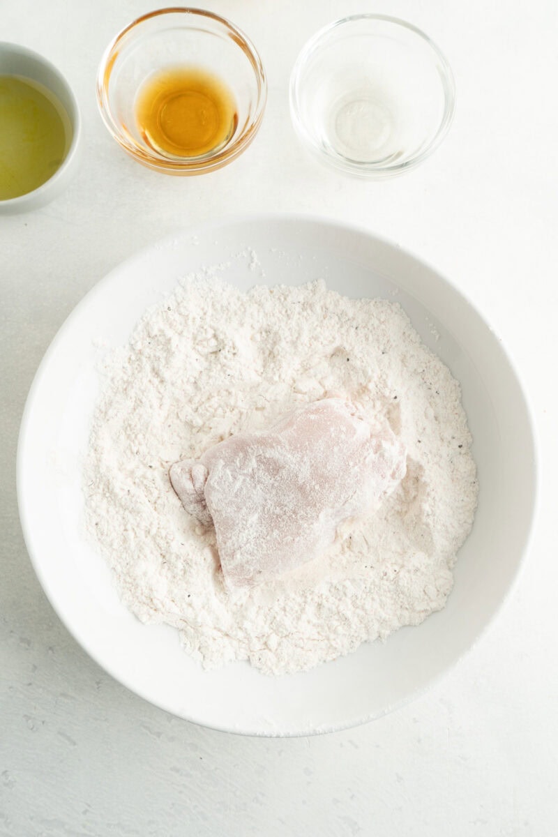 A chicken thigh being dredged in a bowl of seasoned flour.