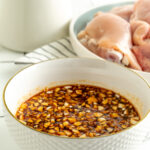 A bowl of honey sriracha sauce next to a plate of raw chicken.