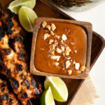 An overhead shot of peanut sauce in a wood bowl with skewers on a plate.