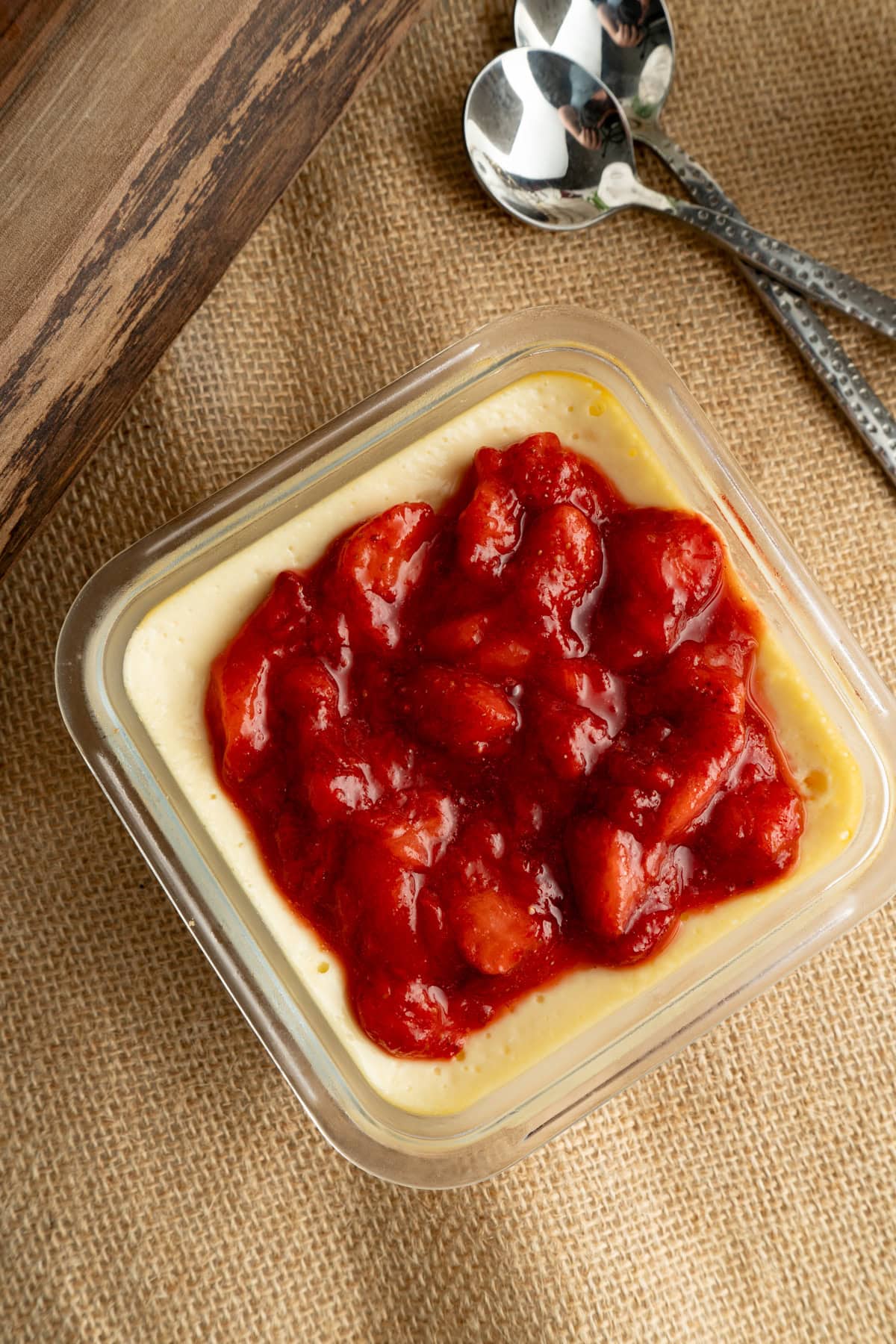 A single strawberry cottage cheese cheesecake cup on burlap with two spoons.