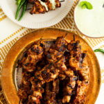 A gold rimmed plate of tandoori chicken skewers on a table.