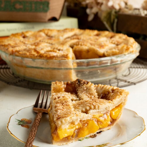 A slice of peach pie and a fork with the whole pie in the background.