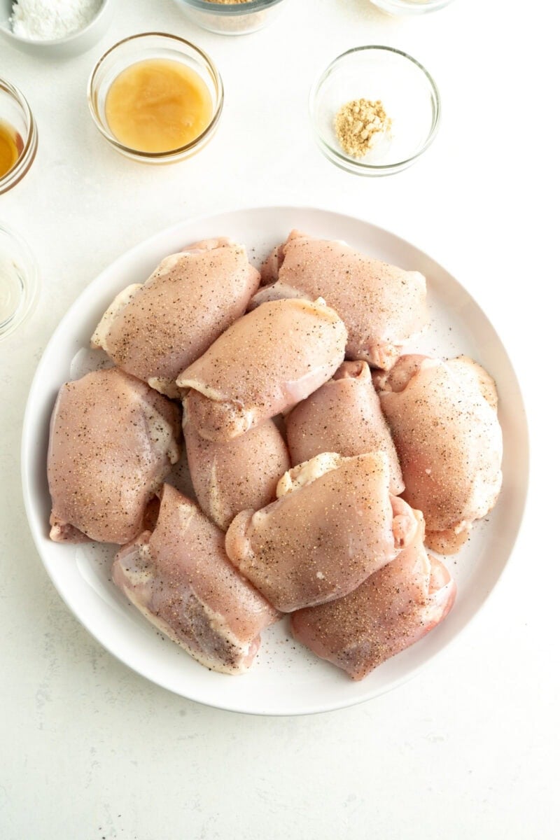 Raw chicken thighs on a plate seasoned with salt and pepper.