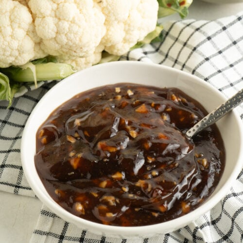 A bowl of Asian BBQ sauce with a spoon on a table with cauliflower.