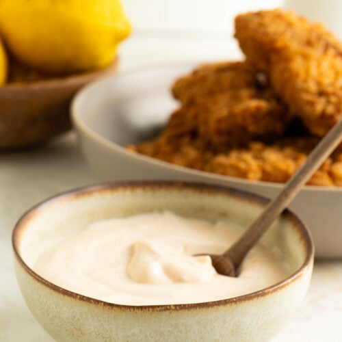A bowl of peri peri greek yogurt dip with a spoon on a table with lemons and chicken fingers.