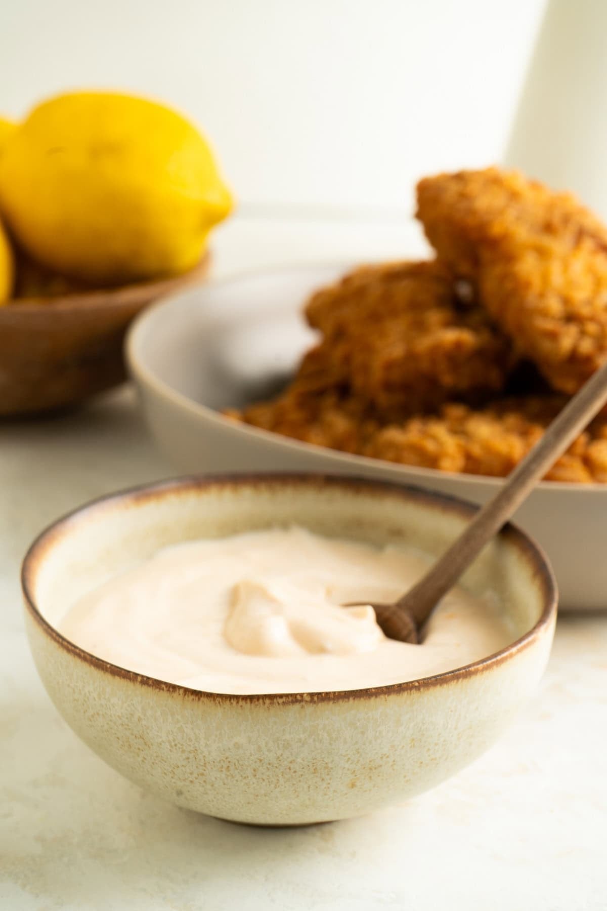 A bowl of peri peri greek yogurt dip with a spoon on a table with lemons and chicken fingers.