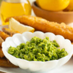A bowl of mushy peas served on a table with fish and chips.