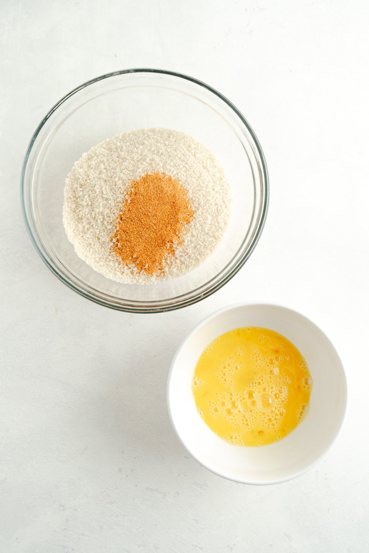 A bowl of bread crumbs with seasonings and a smaller bowl of beaten eggs.