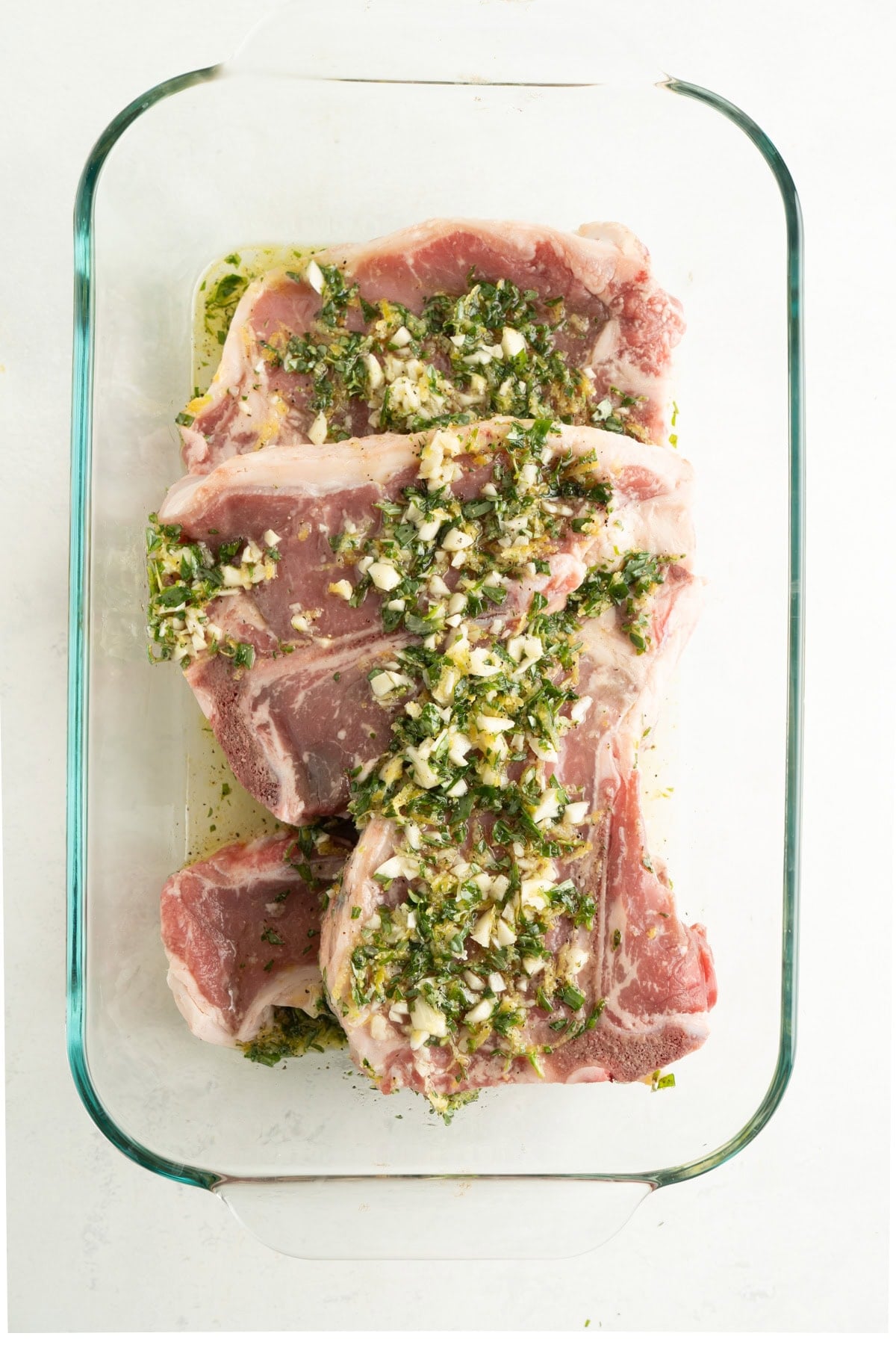 Veal chops in a glass dish with a lemon and herb marinade overtop.