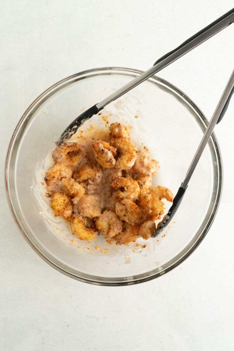 Crispy air fried shrimp being tossed with bang bang sauce in a bowl using tongs.