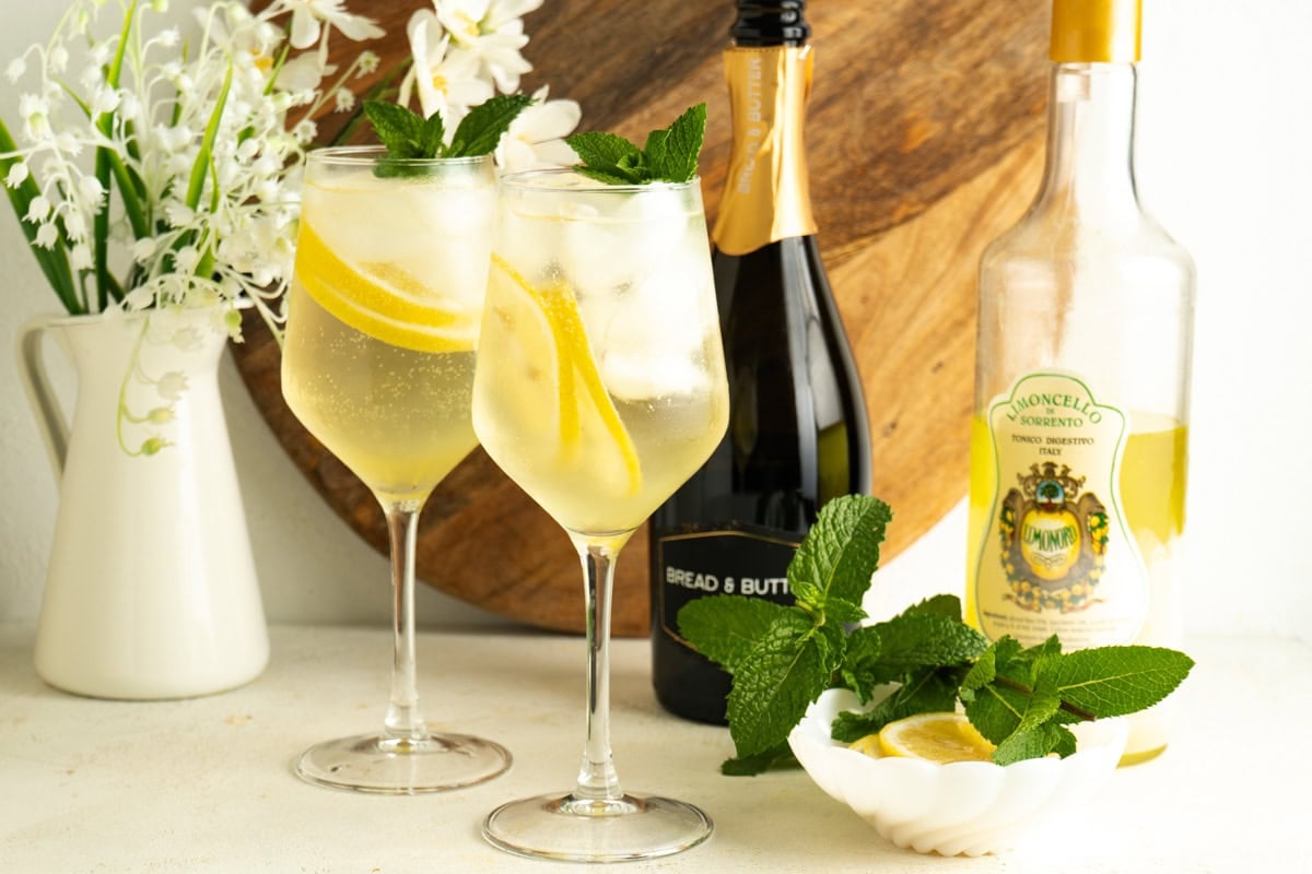 Two wine glasses of limoncello spritz on a table with bottles of prosecco and limoncello.