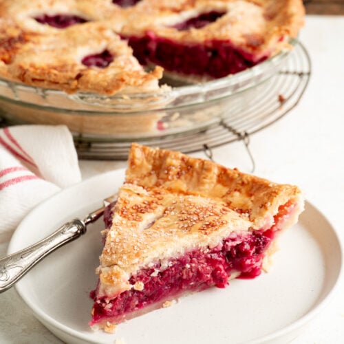A slice of raspberry pie on a plate with a whole pie in the background.