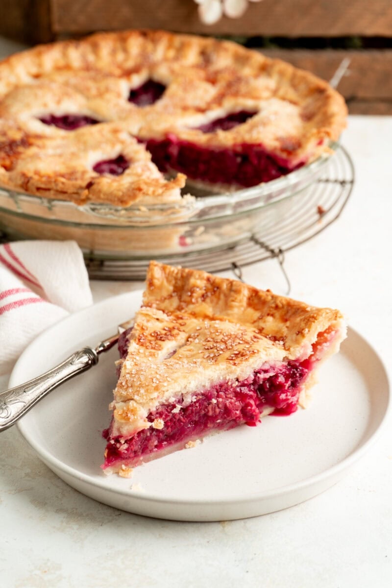 A slice of raspberry pie on a plate with a whole pie in the background.