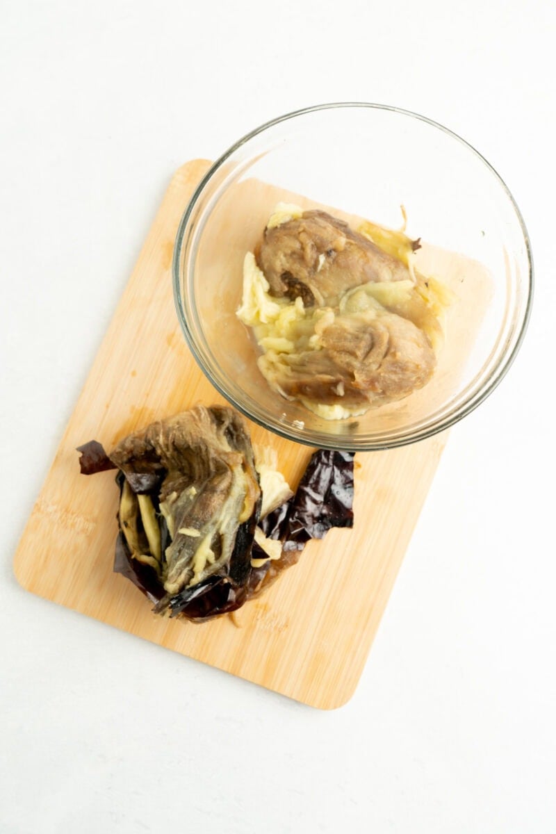 A cooked eggplant in a bowl with the skins removed on the side.